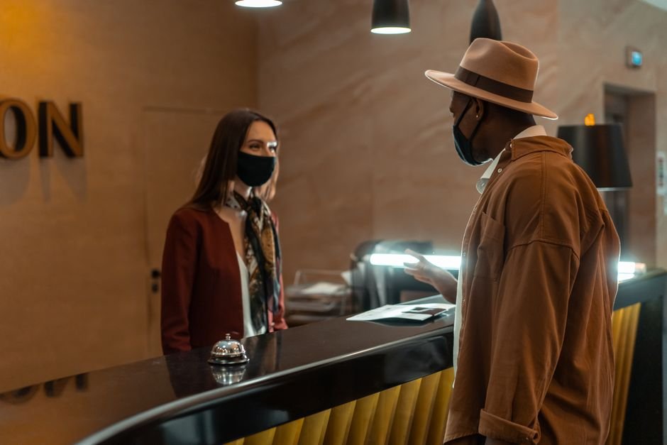 A man at a hotel reception talking to a staff member. Both wear face masks.