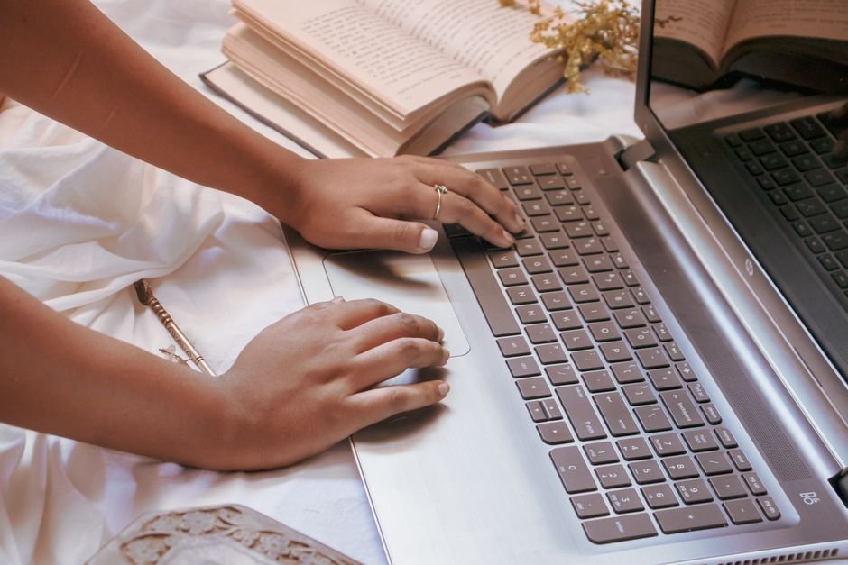 Close-up of a woman typing on a laptop surrounded by open books and a notebook.
