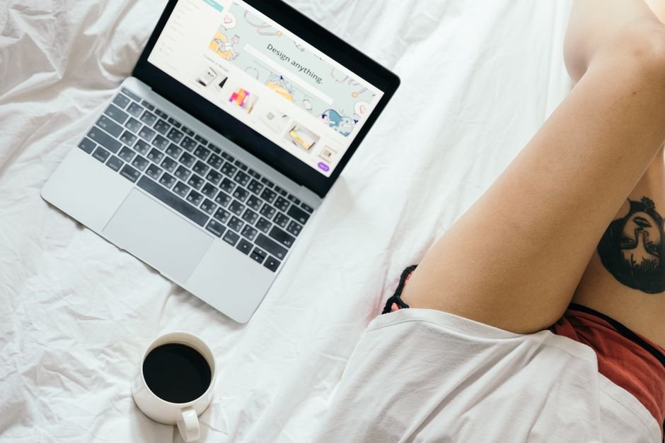 From above of unrecognizable woman with tattoo on leg  browsing netbook and drinking coffee while resting on bed in cozy bedroom