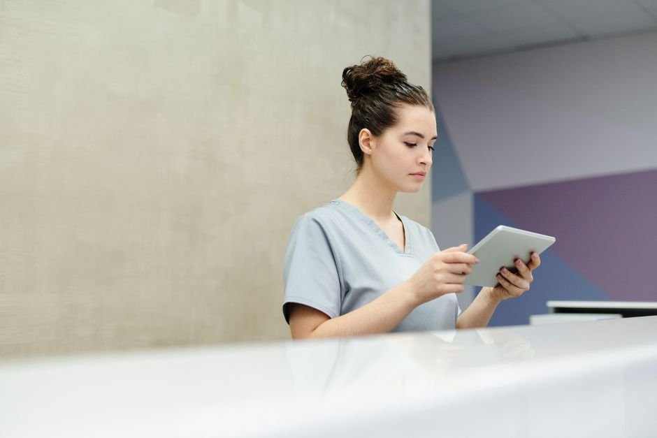 A receptionist in scrubs using a tablet at an office reception desk.